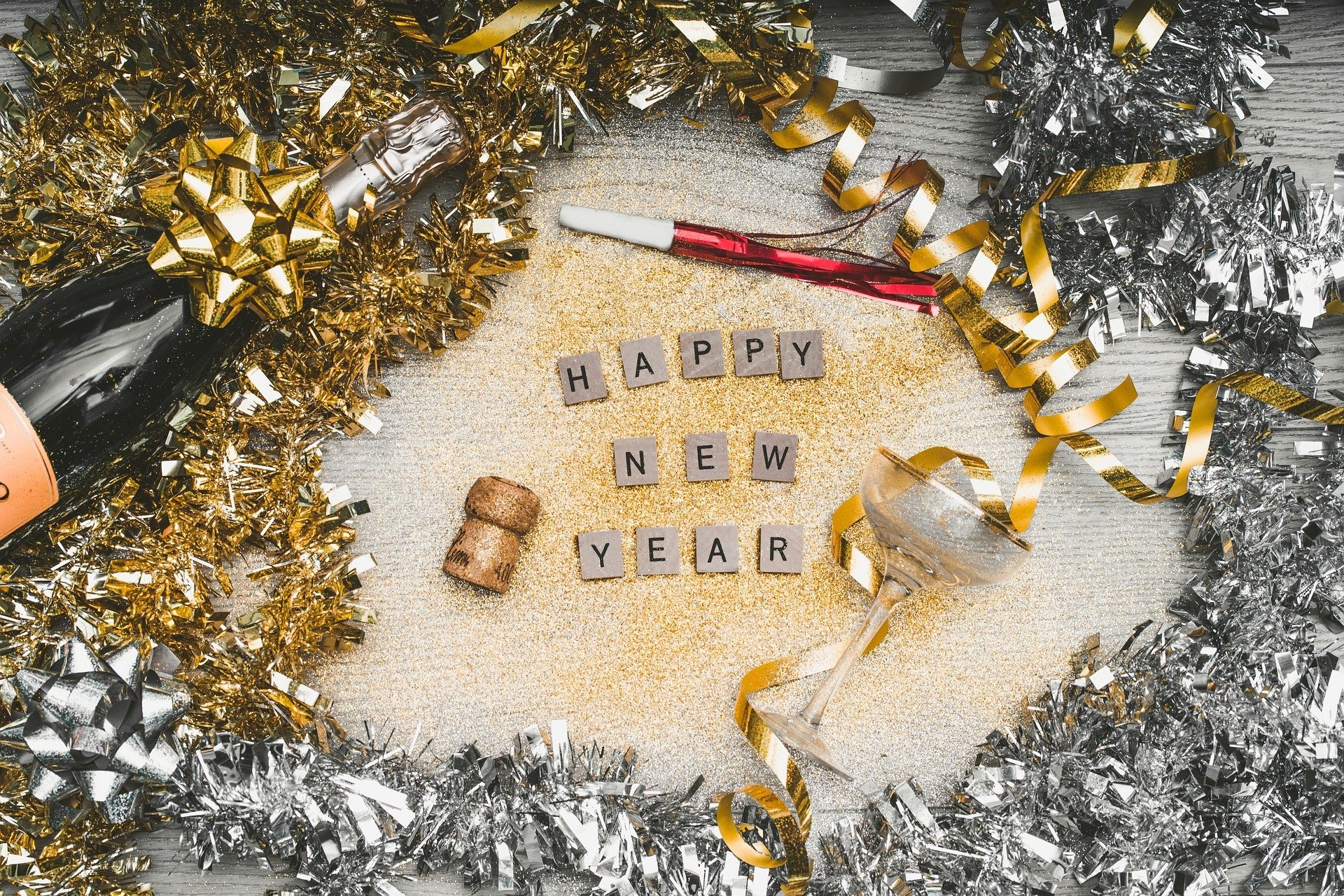 Festive HD PC desktop wallpaper featuring Happy New Year spelled with Scrabble tiles, surrounded by gold and silver holiday decorations, ribbons, and a champagne bottle.
