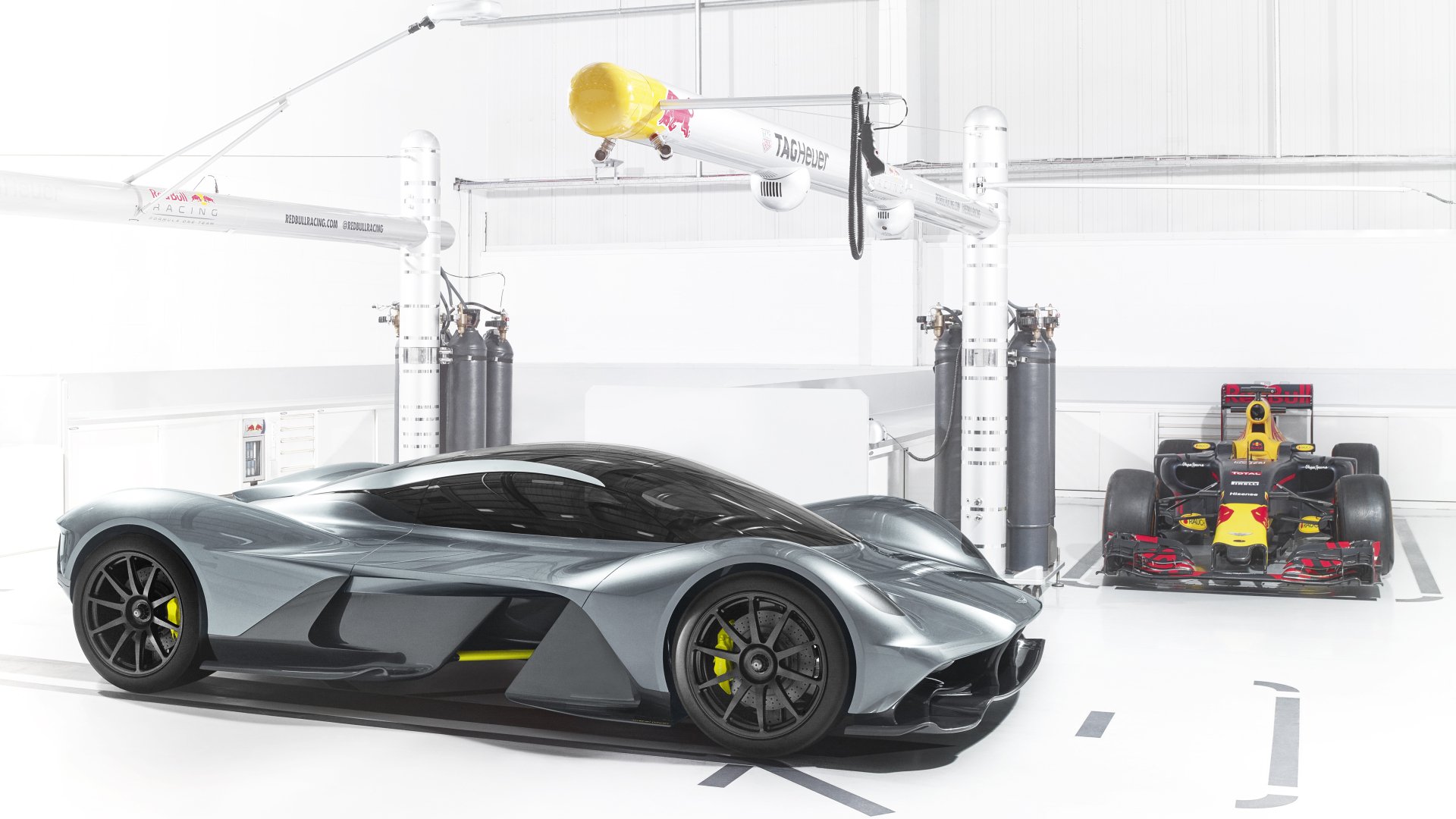 5K Ultra HD PC desktop wallpaper showing an Aston Martin Valkyrie hybrid supercar vehicle in a white garage, sleek metallic body with yellow accents beside a race car.