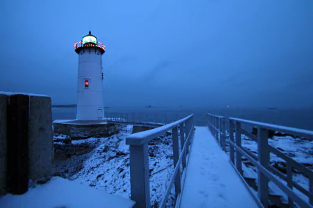 HD wallpaper of a lighthouse with Christmas lights by the ocean, connected by a snowy walkway and bridge under a deep blue sky.
