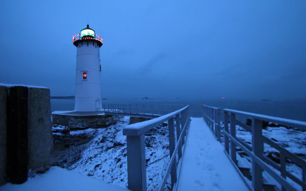 HD wallpaper of a lighthouse with Christmas lights by the ocean, connected by a snowy walkway and bridge under a deep blue sky.