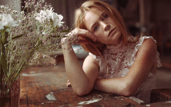 Freckled redhead woman model in a lace top leans on a rustic wooden table beside wildflowers — HD PC desktop wallpaper/background.