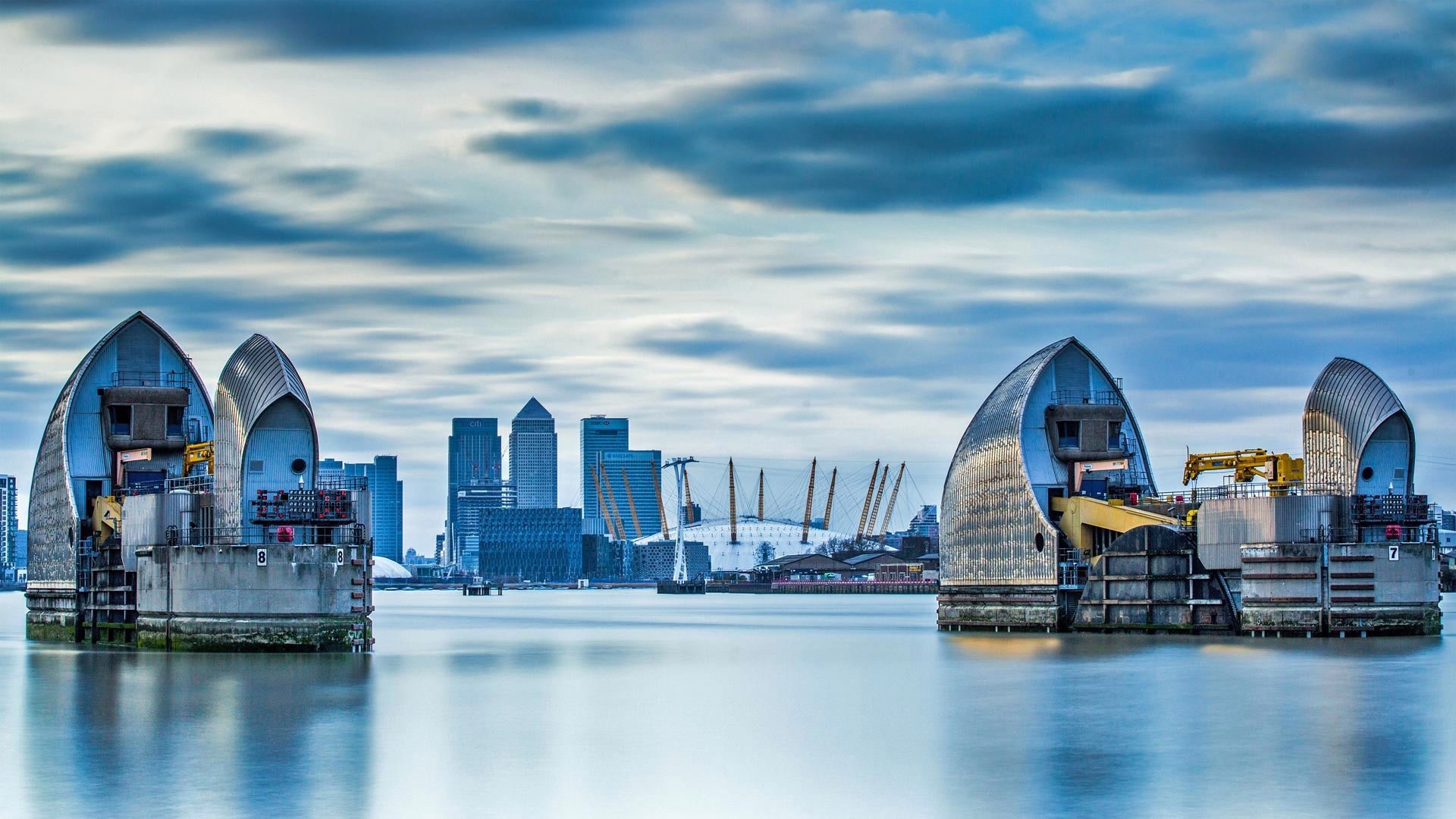 HD PC wallpaper of United Kingdom: London skyline with Thames Barrier floodgates and Canary Wharf skyscrapers reflected on the river beneath a cloudy sky.