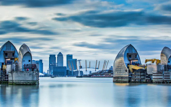 HD PC wallpaper of United Kingdom: London skyline with Thames Barrier floodgates and Canary Wharf skyscrapers reflected on the river beneath a cloudy sky.