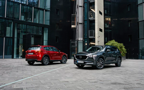 Two Mazda CX-5 SUVs, red and gray, parked in a modern glass-and-concrete plaza, shown as a 2K Quad HD PC desktop wallpaper background.