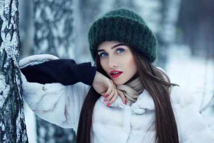 A brunette woman with long hair, blue eyes, and red lipstick stands in a winter forest wearing a green hat and white coat, captured with a depth of field effect. HD desktop wallpaper.