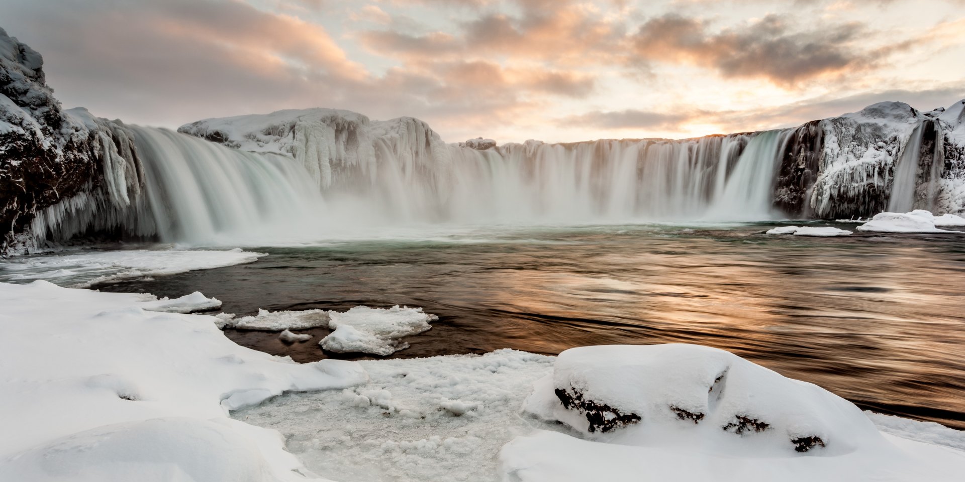 Goðafoss waterfall in winter, surrounded by snow and icy rocks, under a vibrant sunset sky with clouds, captured in stunning 4K Ultra HD quality.