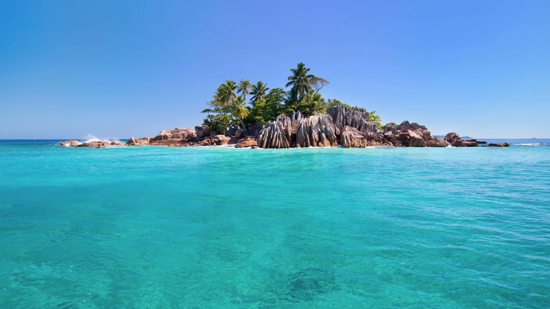 Turquoise tropical sea surrounds a small island in the Seychelles, featuring palm trees and rocky shores under a clear blue sky.