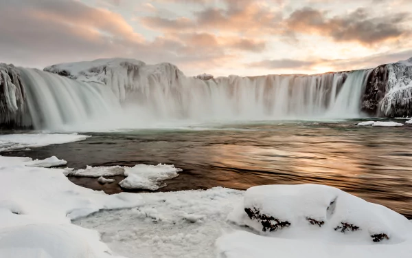 Goðafoss waterfall in winter, surrounded by snow and icy rocks, under a vibrant sunset sky with clouds, captured in stunning 4K Ultra HD quality.