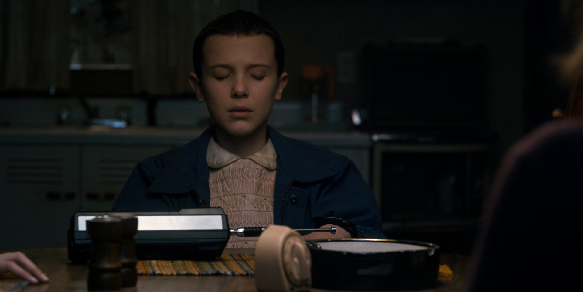 Eleven from Stranger Things, played by Millie Bobby Brown, is shown focusing at a dimly lit table in this HD PC desktop wallpaper from the TV show.