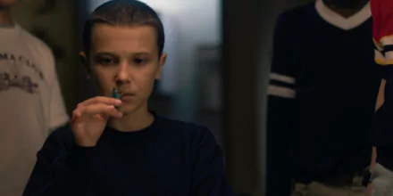 Close-up of Eleven (Stranger Things) concentrating, holding a small object — moody TV show portrait styled as a 2K Quad HD PC desktop wallpaper.