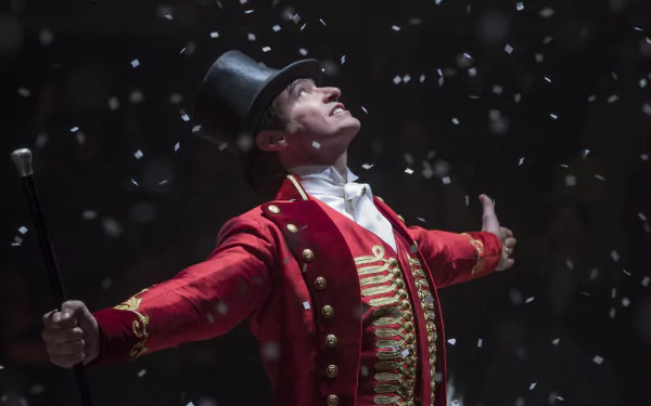 A performer from The Greatest Showman, in a red ringmaster jacket and top hat, exulting amid falling confetti. HD wallpaper.