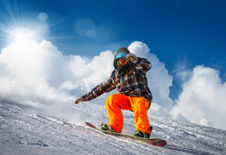 A snowboarder in bright orange pants and plaid jacket descends a snowy slope under a bright sun and blue sky with clouds, captured in 4K Ultra HD.