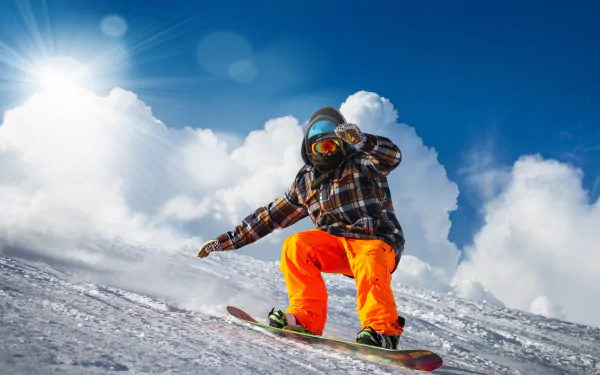 A snowboarder in bright orange pants and plaid jacket descends a snowy slope under a bright sun and blue sky with clouds, captured in 4K Ultra HD.