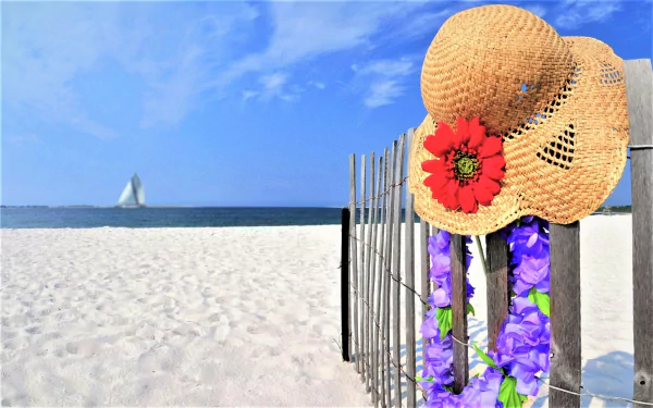 Straw hat adorned with a red flower and purple garland hanging on a beach fence with the ocean and sailboat in the background. HD summer wallpaper.