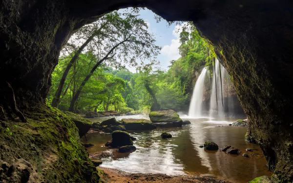 HD desktop wallpaper of a serene lake and waterfall seen through the mouth of a cave, surrounded by lush green trees and nature.