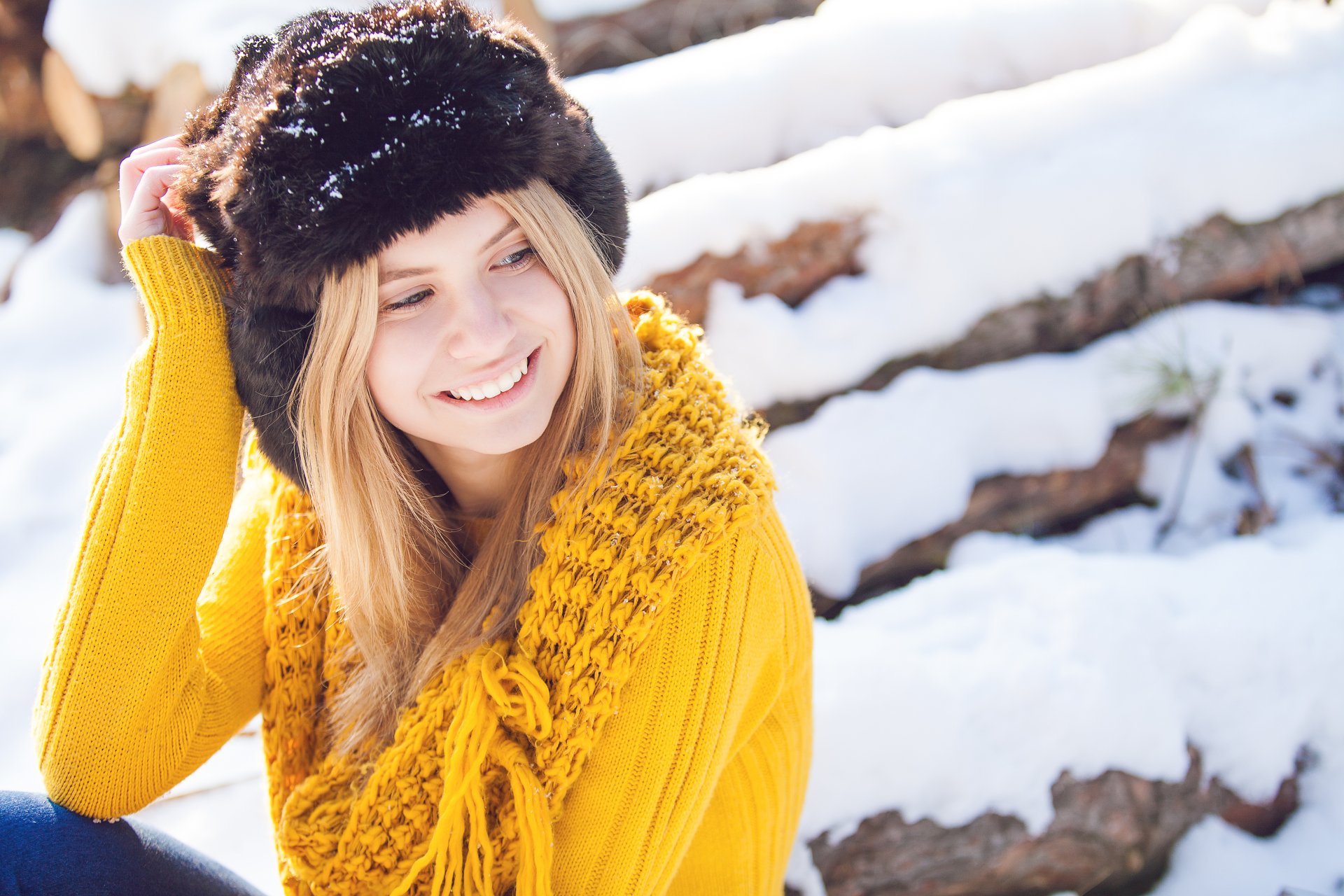 8K Ultra HD PC desktop wallpaper: smiling blonde model in a fur hat and chunky yellow sweater, posing against snowy logs in a bright winter scene.