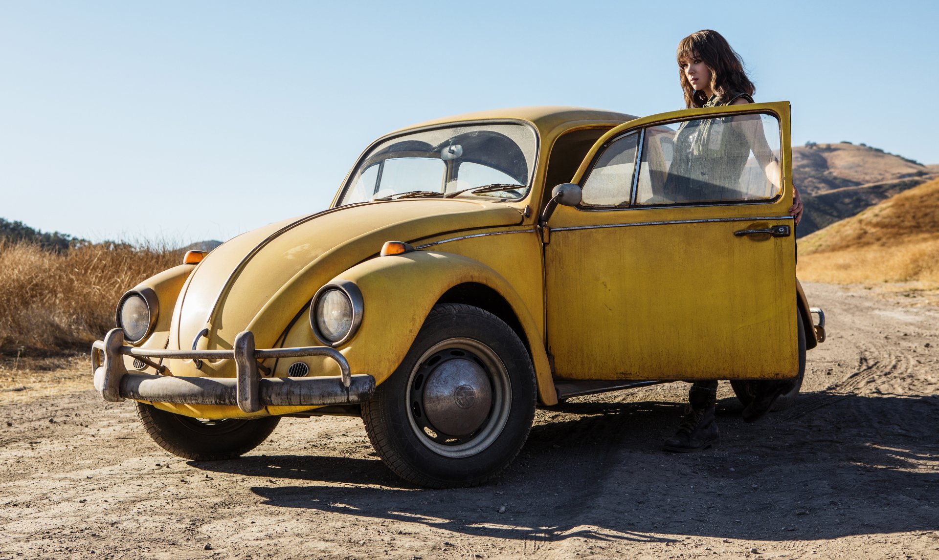 Hailee Steinfeld as a brunette actress stands beside a yellow Volkswagen Beetle, styled as Bumblebee from the Transformers movie, in a 4K Ultra HD desktop wallpaper.