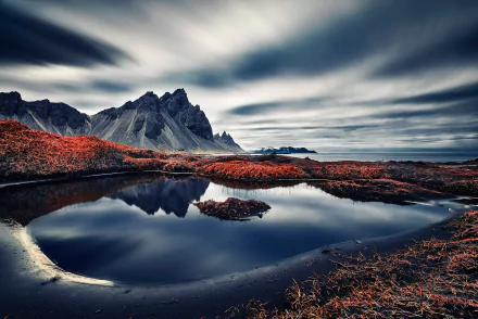 HD desktop wallpaper featuring Vestrahorn in Iceland with a stunning reflection on a calm body of water, capturing the serene horizon, rugged mountain peaks, and dramatic clouds over the tranquil ocean.