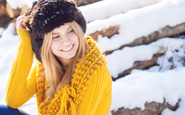 8K Ultra HD PC desktop wallpaper: smiling blonde model in a fur hat and chunky yellow sweater, posing against snowy logs in a bright winter scene.