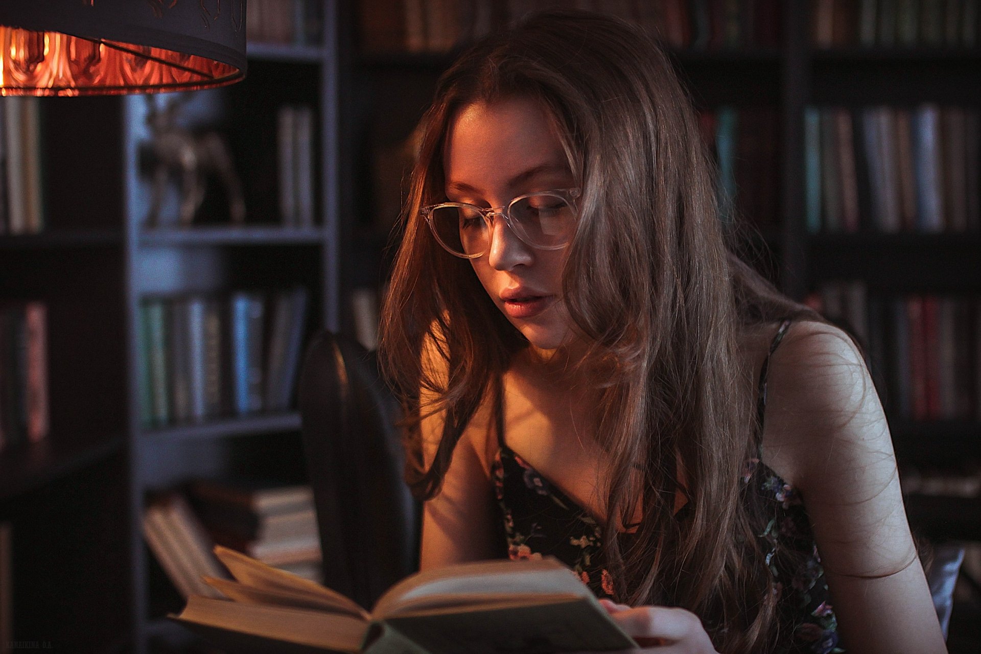 Brunette woman with long hair and glasses reading a book in a dimly lit room, captured as an HD PC desktop wallpaper with a bookshelf background.