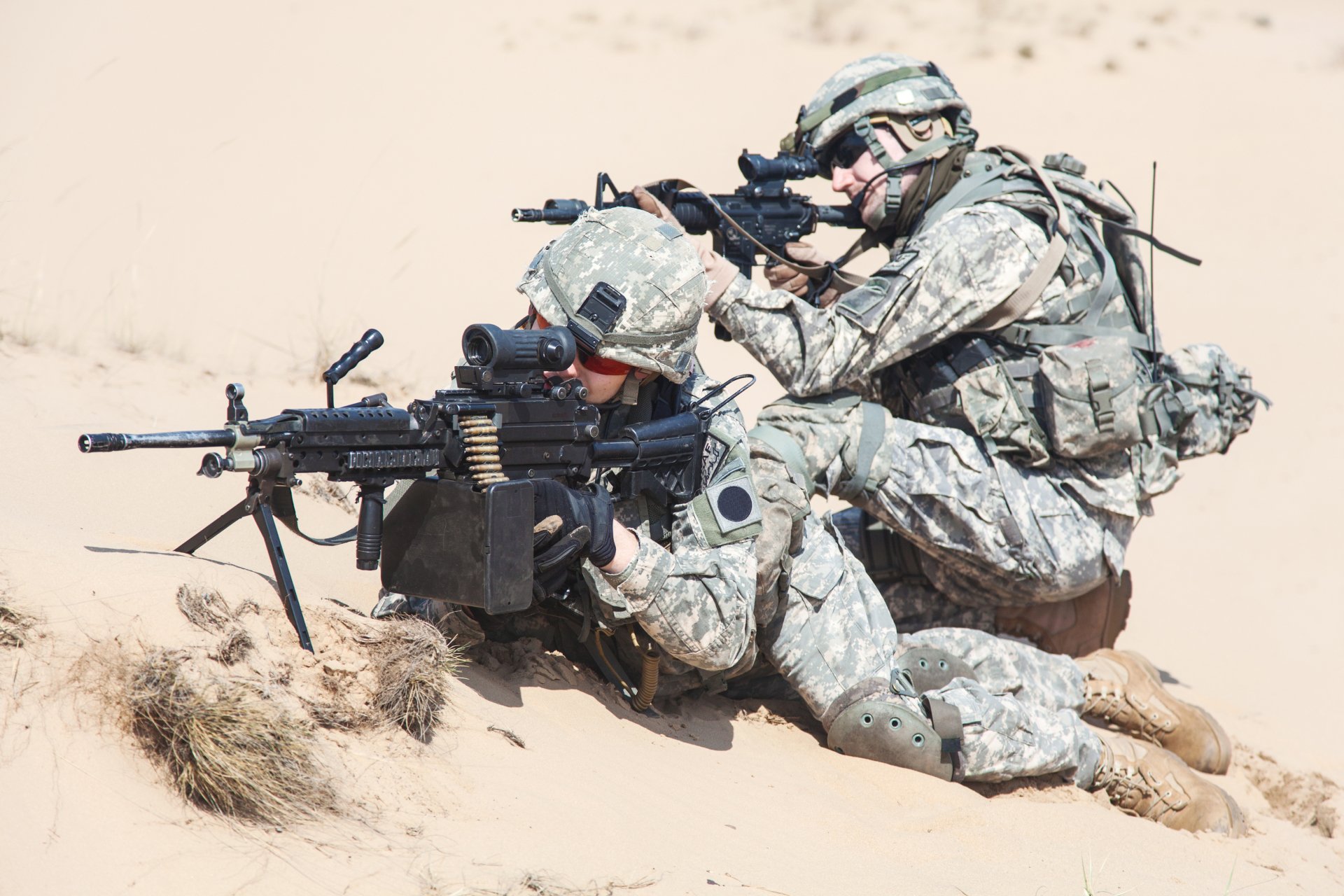 Two soldiers in military gear operate a machine gun while positioned on sand, captured in sharp detail for a 4K Ultra HD PC desktop wallpaper.