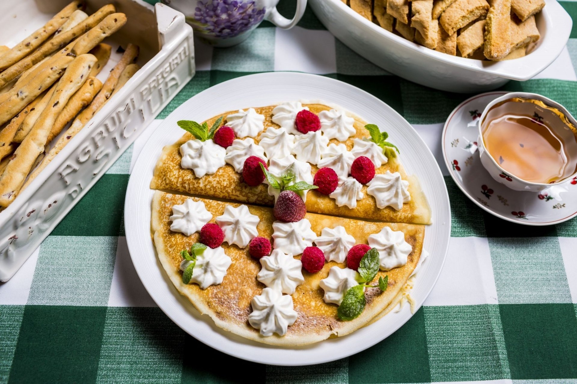 HD PC desktop wallpaper and background — still life of a crêpe topped with cream, raspberries and mint on a white plate, with a teacup and biscuits on a green check tablecloth.