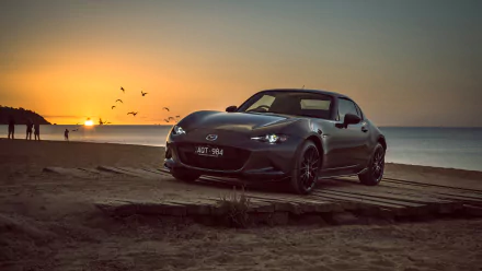 A Mazda MX-5 sports car parked on a beach at sunrise, captured in 4K Ultra HD, with the ocean and birds flying in the background.