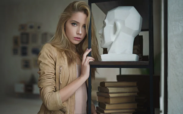 Blonde model Ksenia Kokoreva poses with a focused depth of field, standing beside a geometric white sculpture and stacked books in this HD desktop wallpaper.