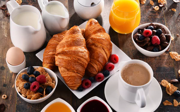 A 4K Ultra HD still life breakfast featuring croissants, muesli with blueberries and raspberries, coffee with milk, orange juice, and an egg on a rustic wooden table.