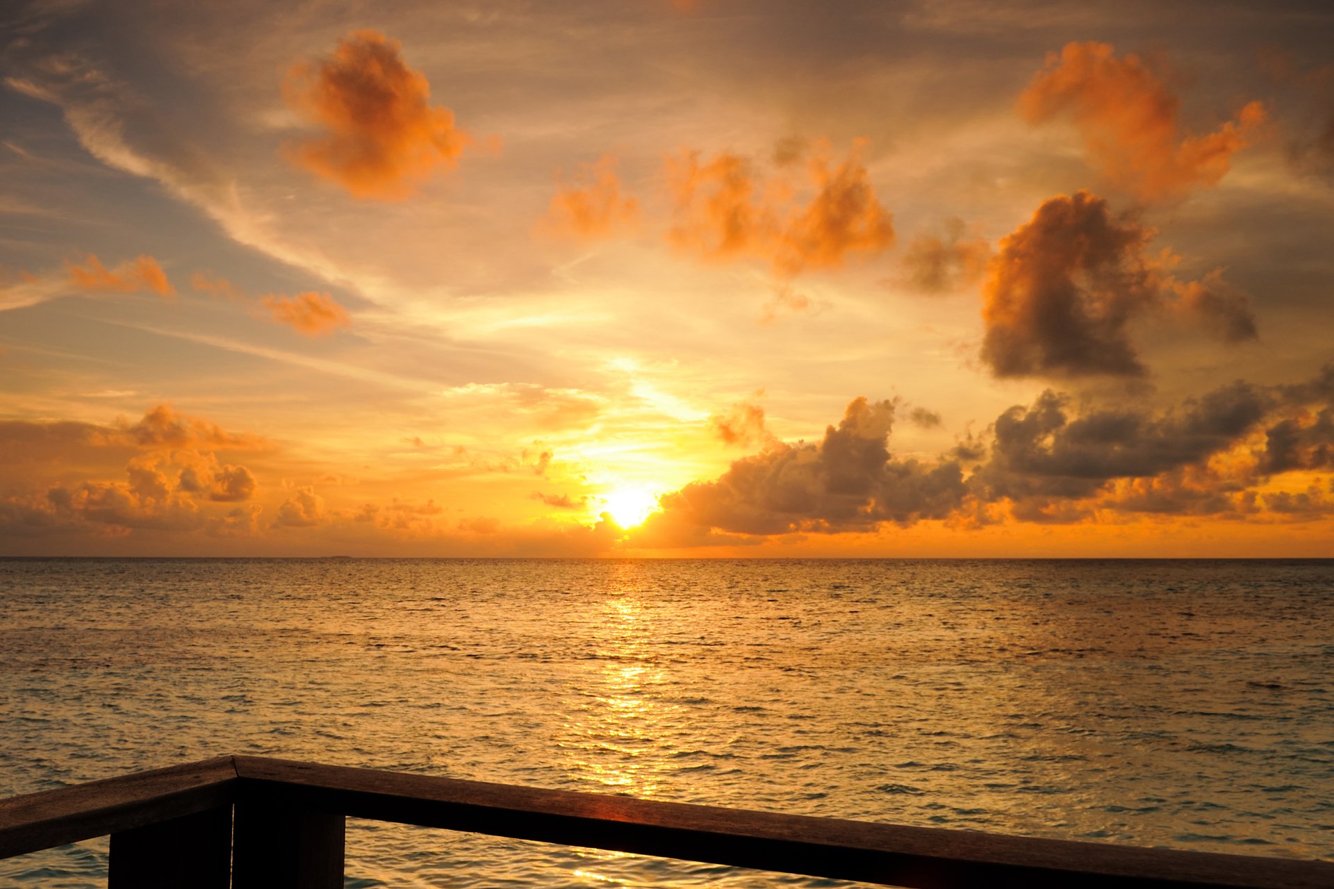 2K Quad HD PC desktop wallpaper and background: Maldives sunset over the horizon, golden sky and clouds reflecting on calm ocean with a wooden railing in the foreground.