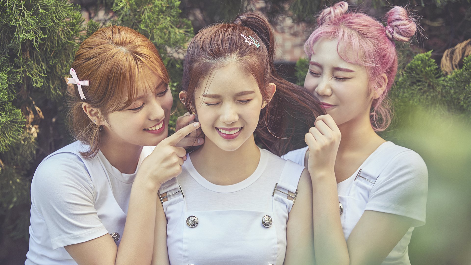 Three smiling Korean girls from the K-pop girl band Momoland with blonde, brunette, and pink hair, wearing casual white outfits in a bright outdoor setting.