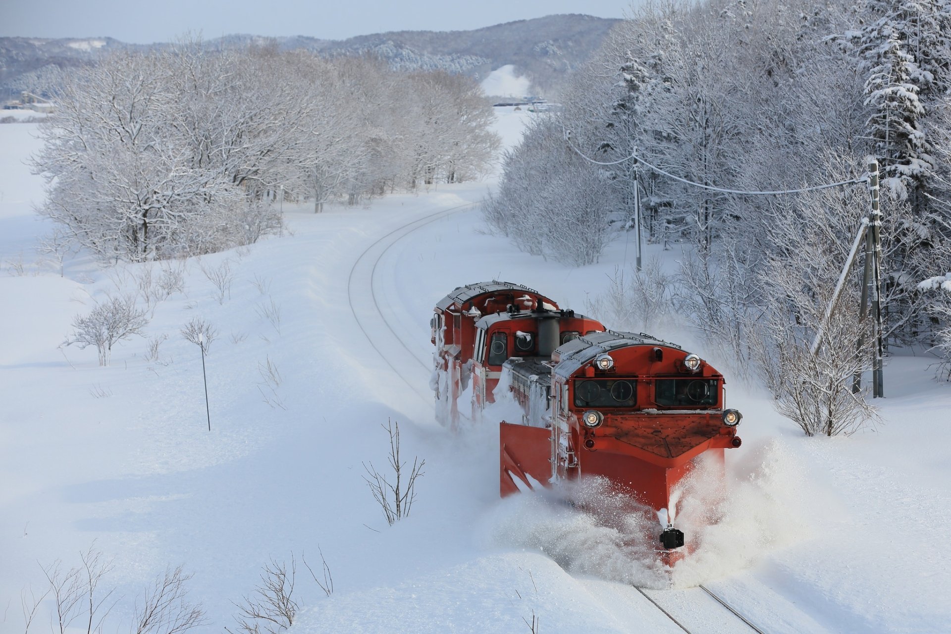 A red locomotive powers through heavy snow in a serene winter landscape, surrounded by snow-covered trees, as captured in this HD desktop wallpaper background.