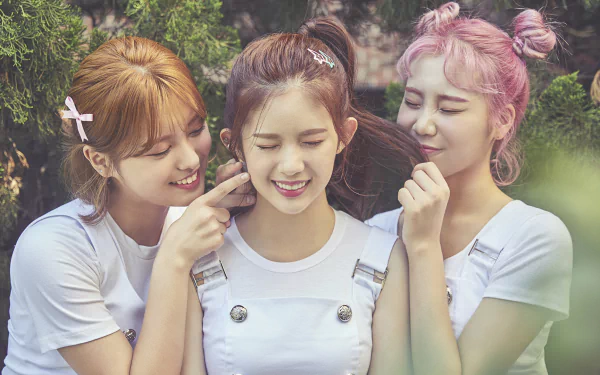 Three smiling Korean girls from the K-pop girl band Momoland with blonde, brunette, and pink hair, wearing casual white outfits in a bright outdoor setting.