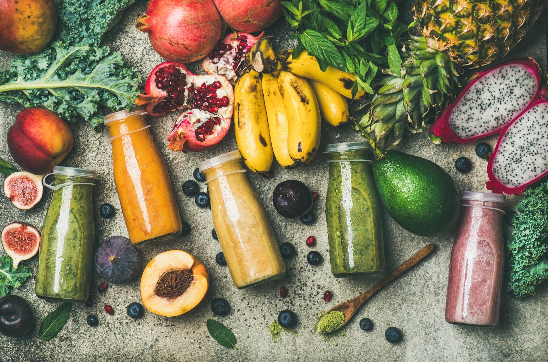 8K Ultra HD still life desktop wallpaper featuring vibrant fruit smoothies in glass bottles surrounded by bananas, pineapple, pomegranate, avocado, figs, and leafy greens.