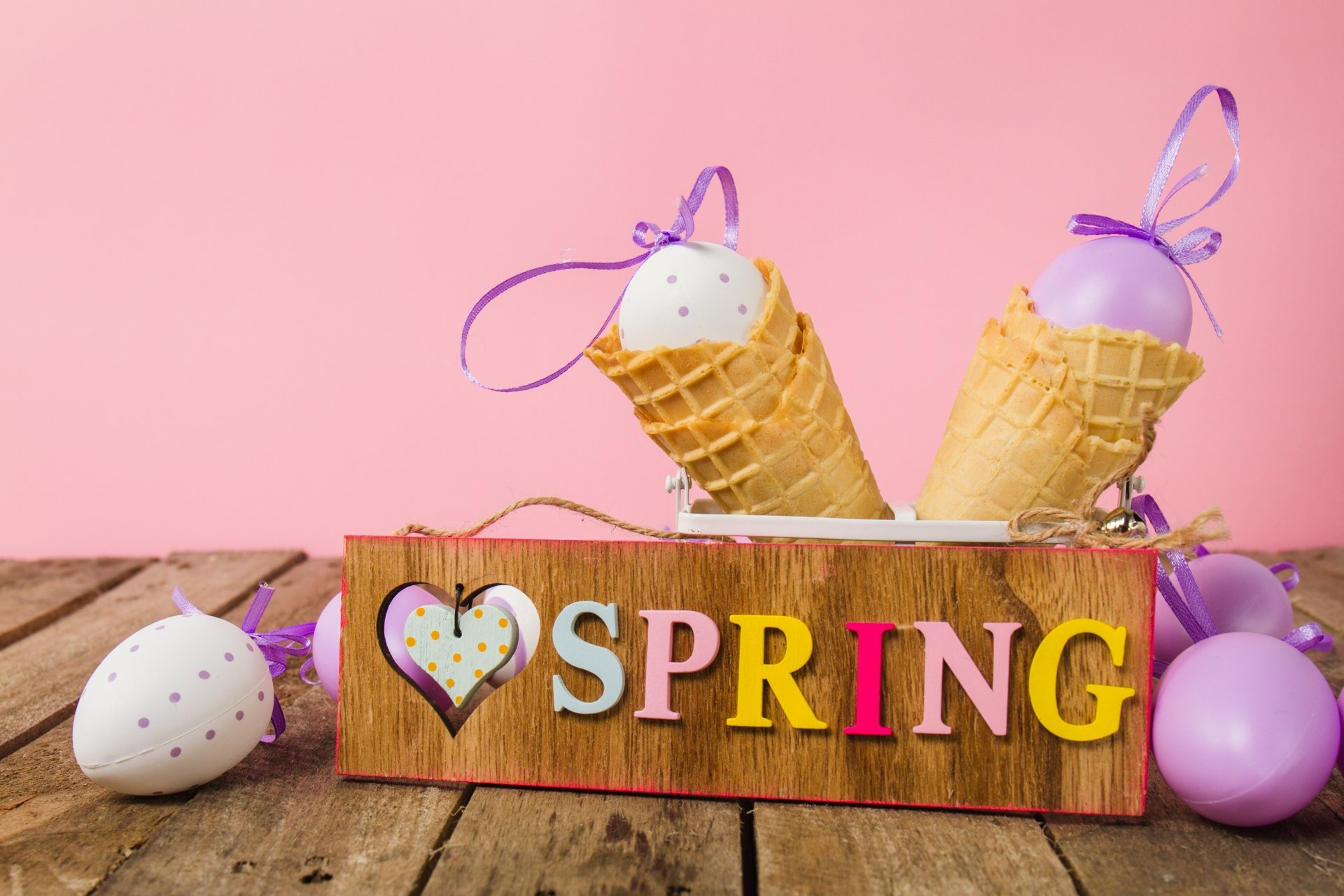 A still life 4K Ultra HD wallpaper featuring pastel eggs in waffle cones with purple ribbons, set against a pink background and a wooden SPRING sign.