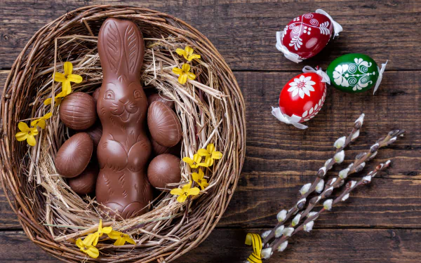 4K Ultra HD still life of a chocolate Easter bunny and eggs nestled in a rustic nest with yellow flowers, alongside painted eggs and pussy willow branches on a wooden background.