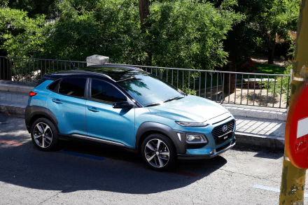 Blue Hyundai Hyundai Kona SUV parked by a tree-lined sidewalk, shown as a 2K Quad HD PC desktop wallpaper/background.