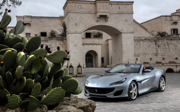 A silver Ferrari Portofino convertible parked in front of an historic stone building with cactus plants nearby, captured in stunning 4K Ultra HD detail.