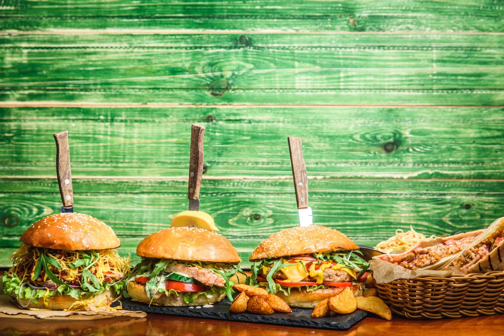 5K Ultra HD PC desktop wallpaper background of food: three gourmet burgers with fries and a bread basket on a rustic green wooden plank backdrop.