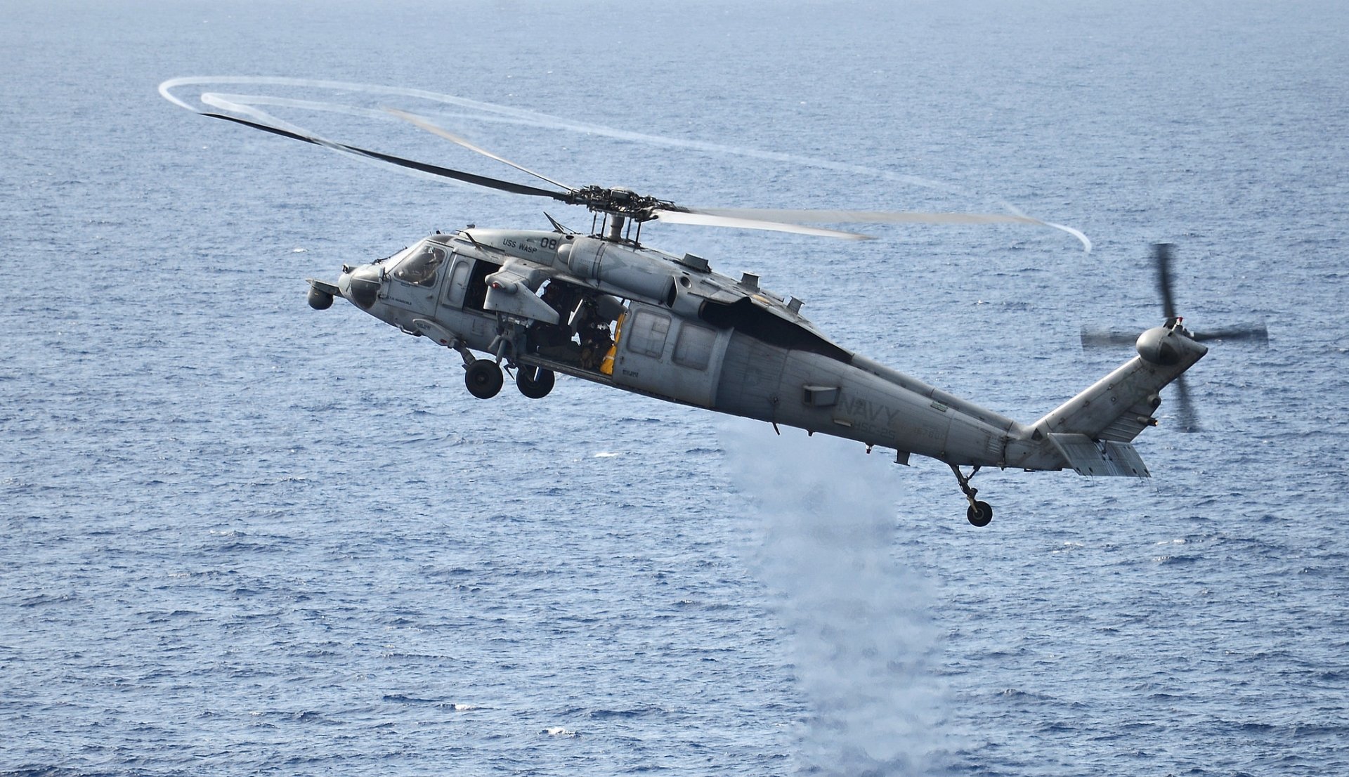 HD desktop wallpaper showing a military Sikorsky SH-60 Seahawk helicopter aircraft banking over the ocean with rotor wake visible.