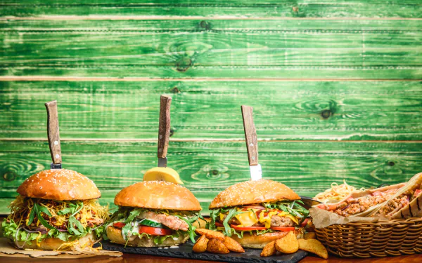 5K Ultra HD PC desktop wallpaper background of food: three gourmet burgers with fries and a bread basket on a rustic green wooden plank backdrop.