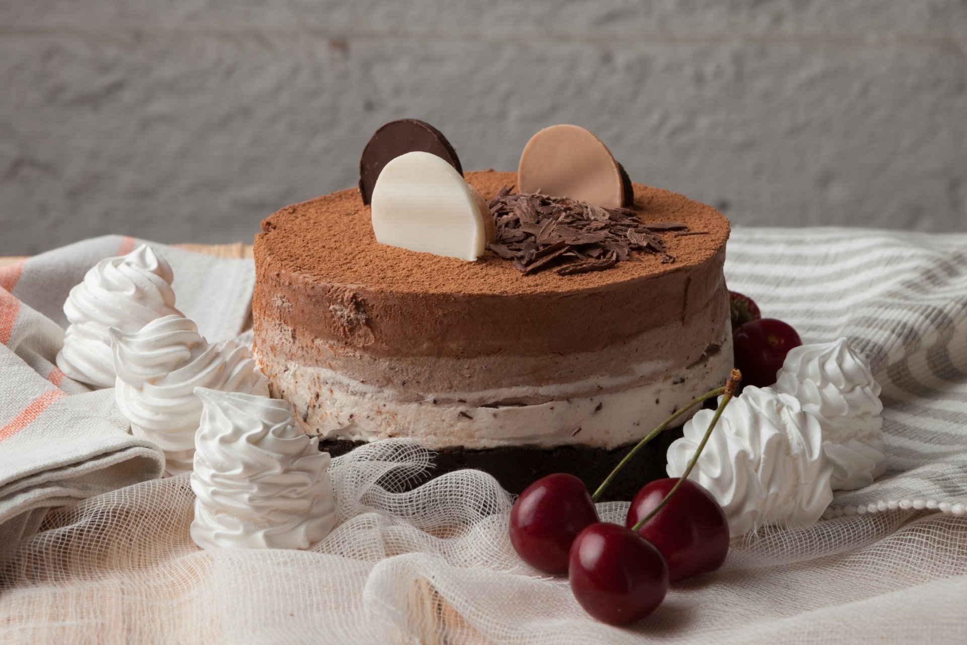 HD desktop wallpaper showcasing a chocolate cake dessert adorned with cherries, meringue swirls, and chocolate decorations on a soft fabric background.