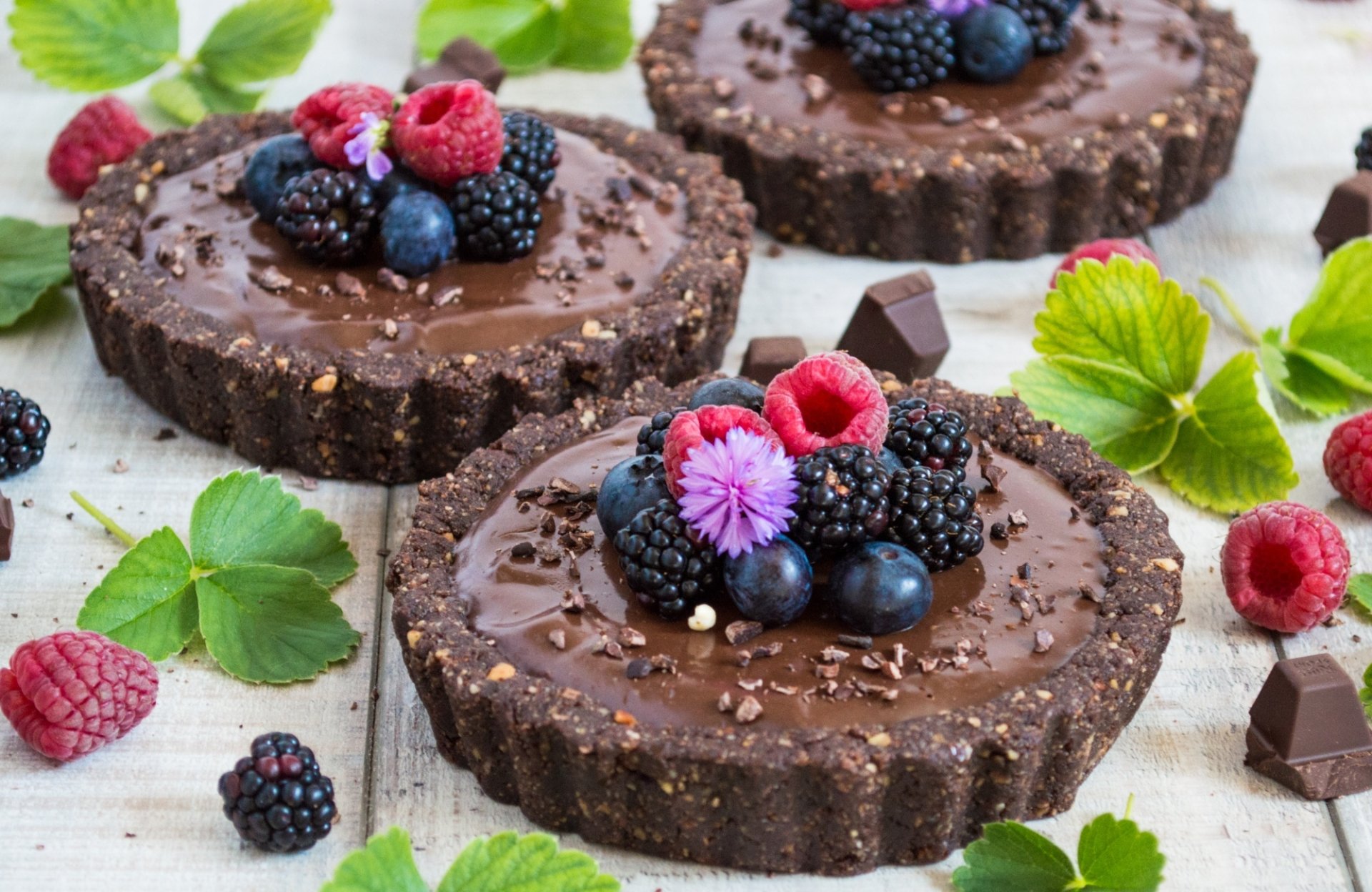 HD desktop wallpaper showing still life of chocolate tarts topped with fresh blueberries, raspberries, and blackberries, surrounded by scattered berries and green leaves.