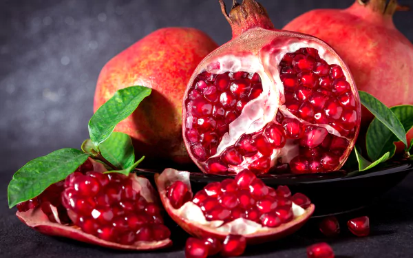 Close-up 4K Ultra HD desktop wallpaper of ripe pomegranates with vibrant red seeds and green leaves on a dark textured background.
