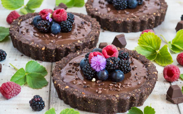 HD desktop wallpaper showing still life of chocolate tarts topped with fresh blueberries, raspberries, and blackberries, surrounded by scattered berries and green leaves.