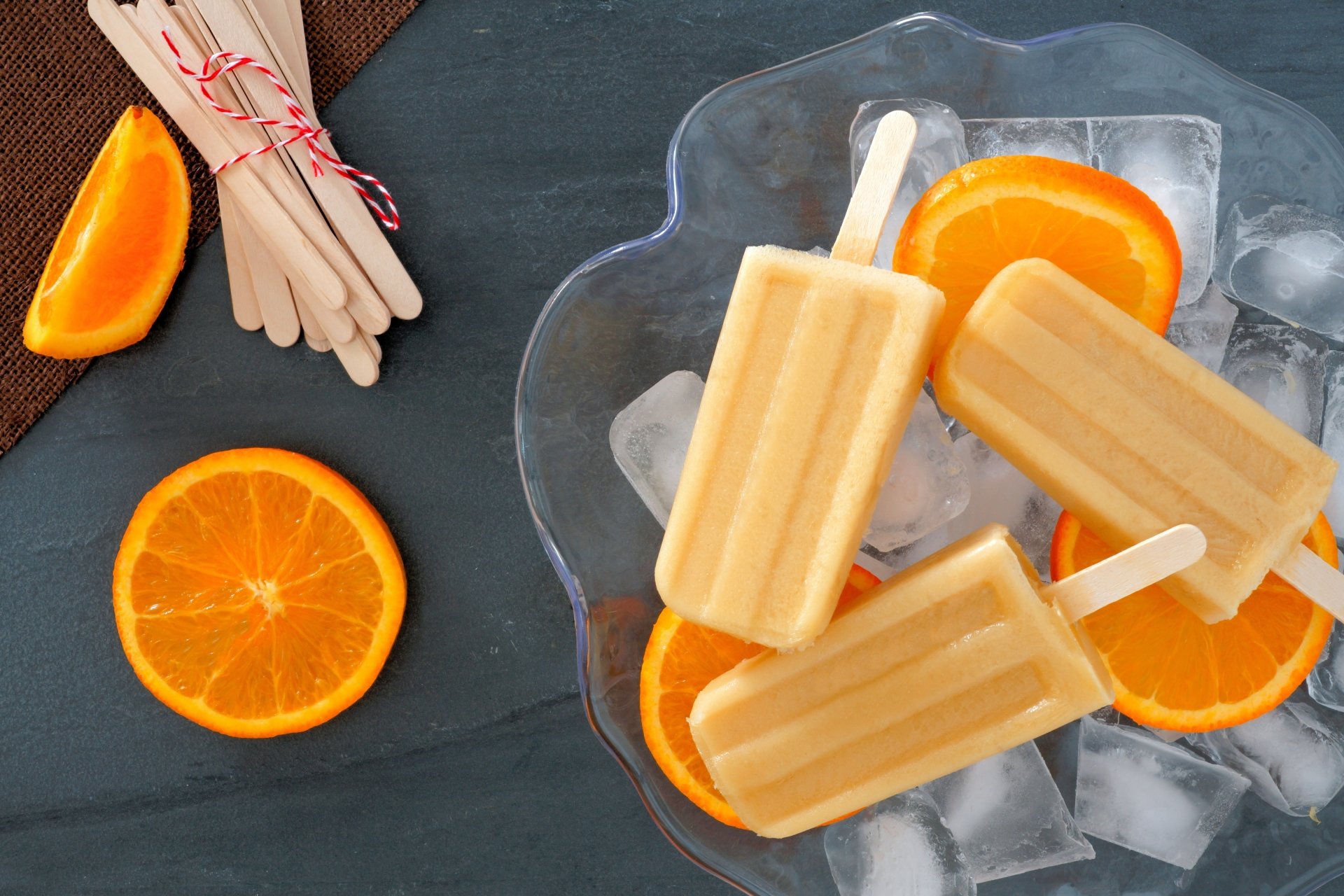 4K Ultra HD still life of orange popsicles on ice cubes with fresh orange slices, set against a dark background for a vibrant food-themed PC desktop wallpaper.
