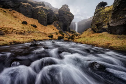HD PC desktop wallpaper and background showing a fast river rushing between mossy cliffs toward a distant waterfall, dramatic nature scenery beneath a cloudy sky.