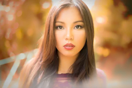 A close-up HD portrait of an Asian brunette model with brown eyes and lipstick, captured with a soft depth of field and warm autumn background.