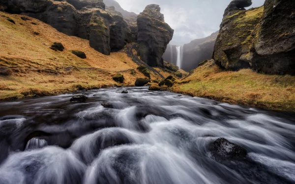 HD PC desktop wallpaper and background showing a fast river rushing between mossy cliffs toward a distant waterfall, dramatic nature scenery beneath a cloudy sky.