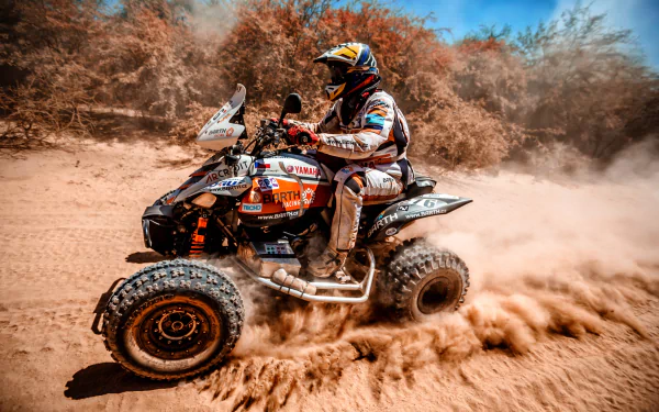 HD PC desktop wallpaper showing a quad vehicle speeding across sandy terrain with a rider in full gear, kicking up dust under a clear blue sky.